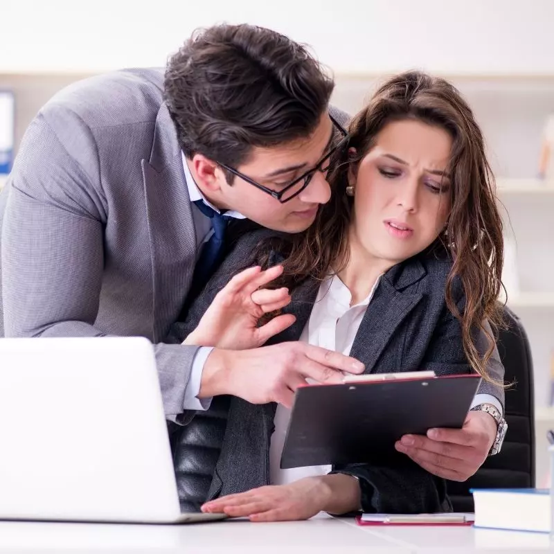 Une femme à son bureau harcelée par un homme qui l’entoure de ses bras pour lui montrer un document avec insistance.